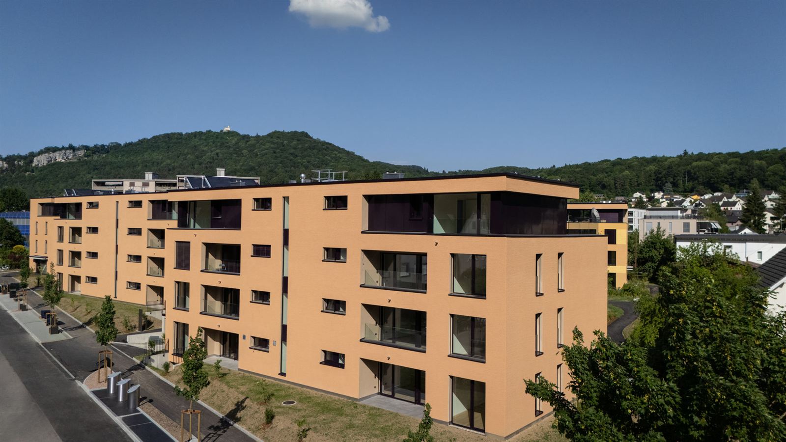 Die Überbauung Im Feldpark von aussen: ein Wohnhaus mit terrakottafarbenem Verputz, Loggien hinter Glasgeländern und einem zurückversetzten Attikageschoss, dahinter der bewaldete Höhenzug über Aarburg.